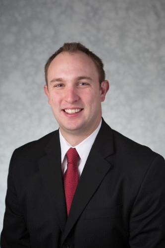 A formal headshot of Jason wearing a black suit and a red tie.